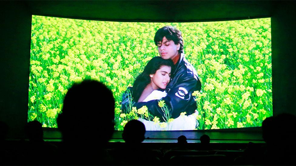 A shot of a scene in a cinema, from Dilwale Dulhania Le Jayenge, a Bollywood fiIm. In the foreground of the image the heads of audience members are just visible in silhouette.