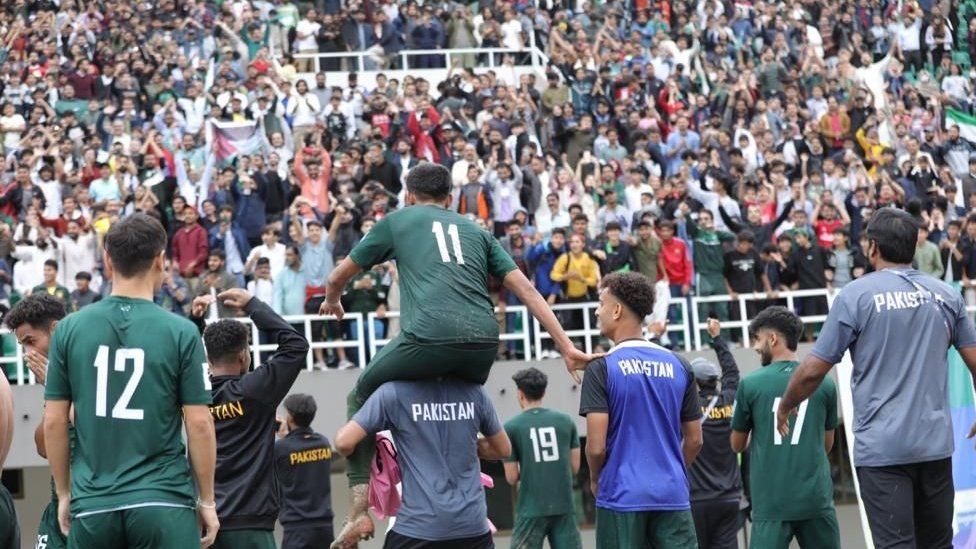 Harun Hamid and other Pakistan players on the pitch celebrating their win against Cambodia last month. Harun is wearing a green football kit with a white number 11 on the back of his shirt and is sat on top of someone else's shoulders facing away from the camera. Other players are around him also facing away from the camera and in front of the players you can see the crowd celebrating.