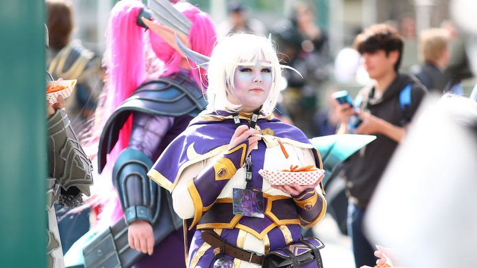 A woman wears a wig and a costume from the game that looks like fantasy armour, while eating a carboard tray of french fries