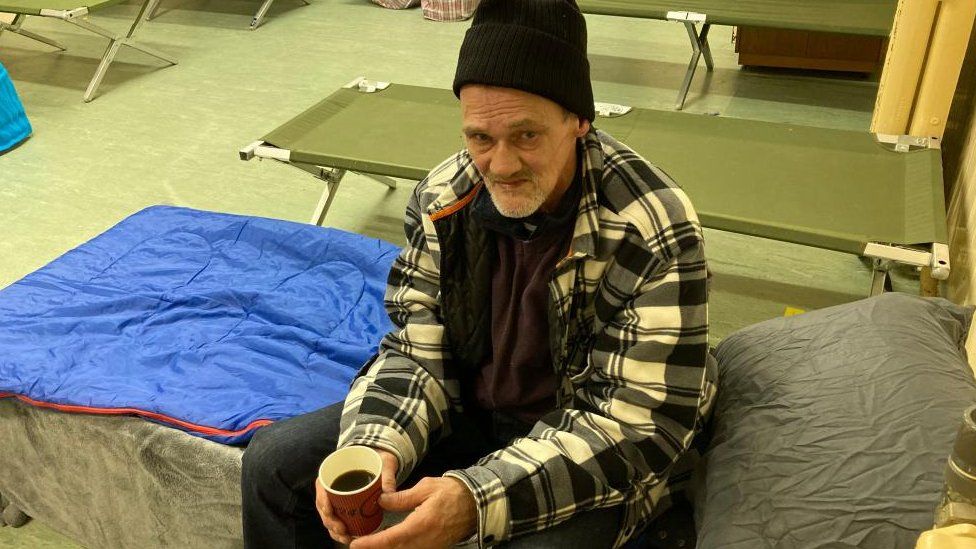 Man wearing a woolly hat sits on a camp bed drinking a hot drink