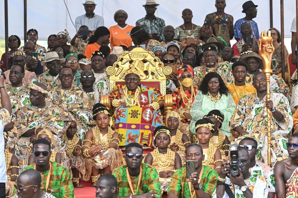 A man on a large gold throne and his entourage watch an event from the stands.