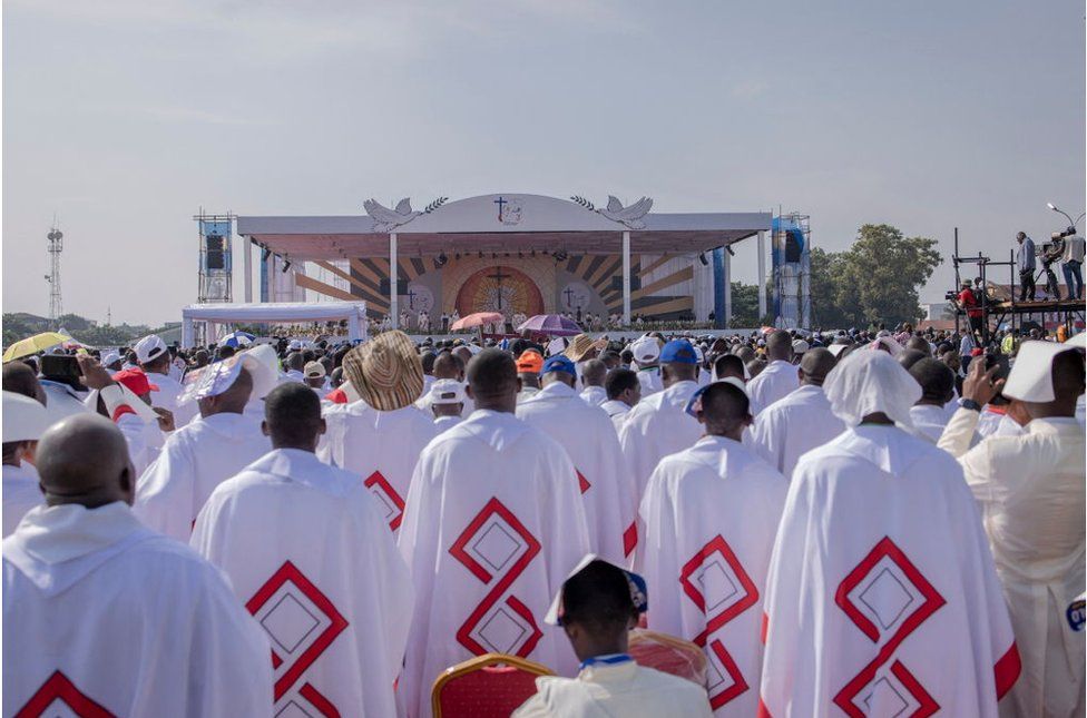 A group of priests wearing their clerical attire facing the stage. They are surrounded by crowds.