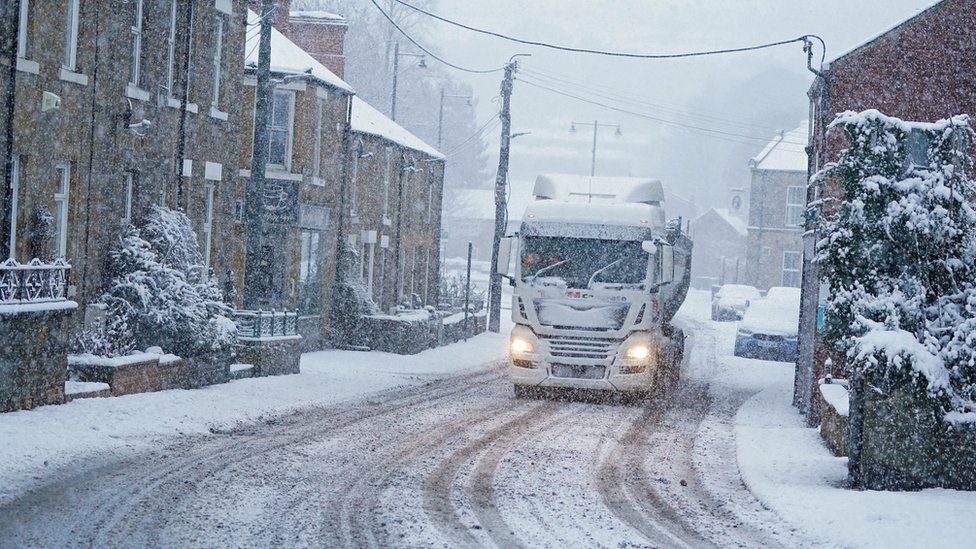 A lorry drives in falling snow past a row of houses