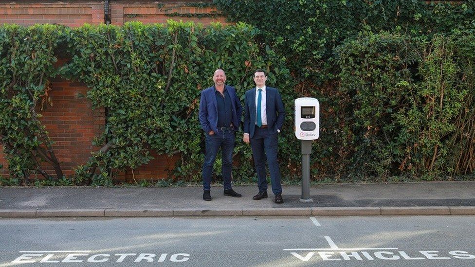 Two men in suits stand on a pavement next to an electric vehicle charging point