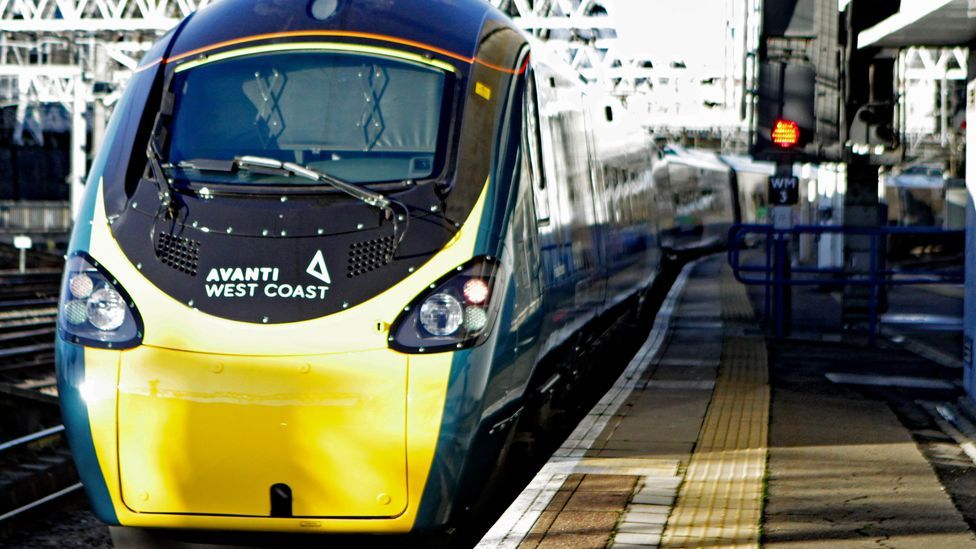 A teal and yellow Avanti West Coast train stands next to a brown and grey platform and alongside a rail signal with a red light