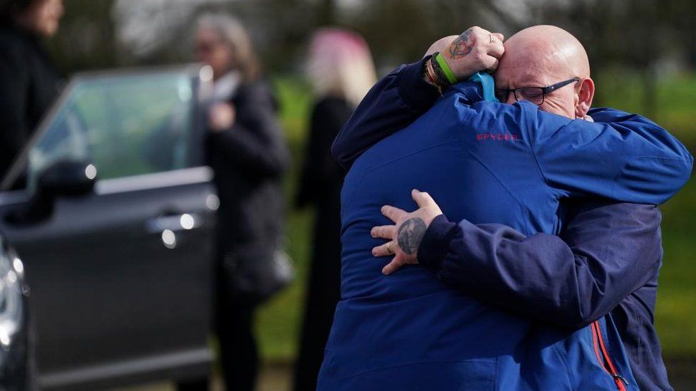 Two mourners hug each other with a hearse in the background