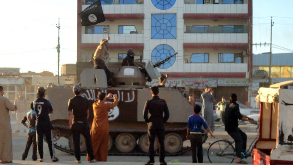 Iraqis stand watching as an armoured vehicle carrying armed militants holding up an Islamic jihadist flag drives past in Falluja on 20 March 2014
