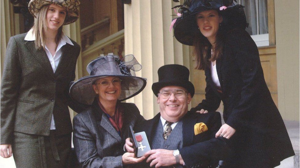 James holding his MBE medal with his wife and daughters by his side