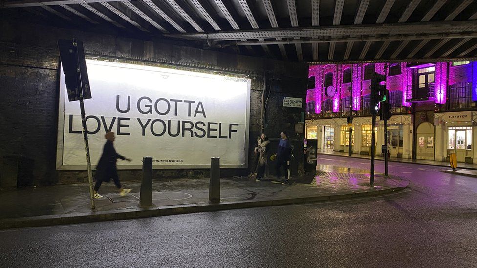 Pedestrians passing an illuminated billboard reading 'U Gotta Love Yourself' beneath the railway bridge on Castlehaven Road at night.