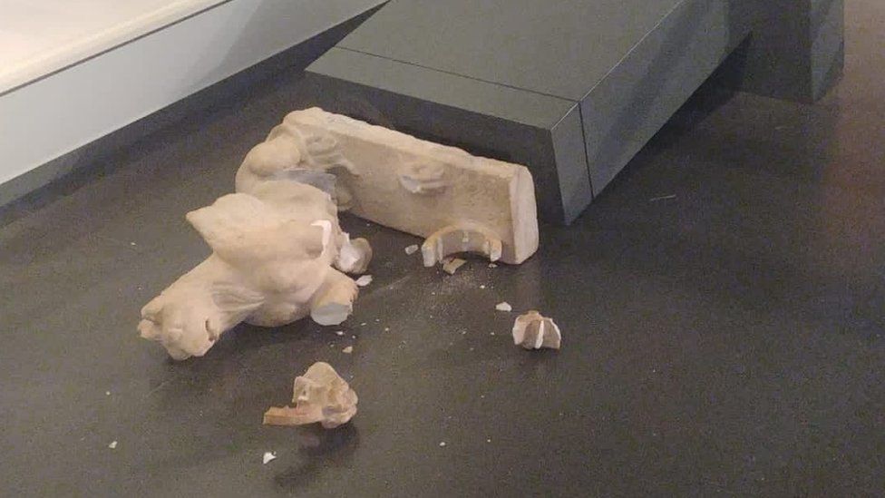 White statue lying smashed on the floor, with the plinth it was standing on knocked over behind it