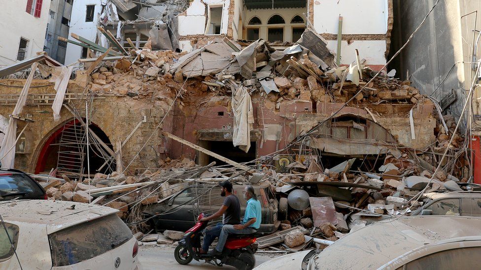 Men ride a motorcycle past a damaged building