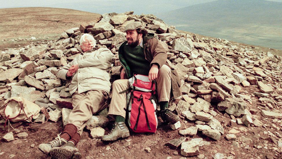 Alfred Wainwright reclining against the summit cairn during filming at Pen-y-ghent in the Yorkshire Dales, alongside presenter Eric Robson