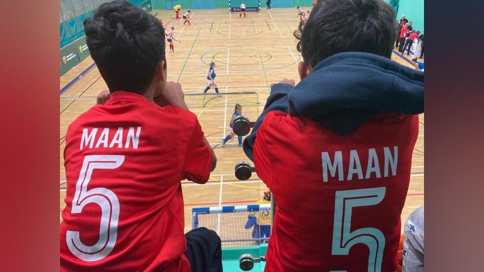 Two boys wearing their mothers' hockey numbers, watching her play