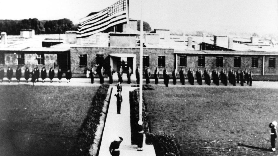 An old black and white image of soldiers on parade in front of a single-storey flat-roofed building with an American flag flying from the flagpole.