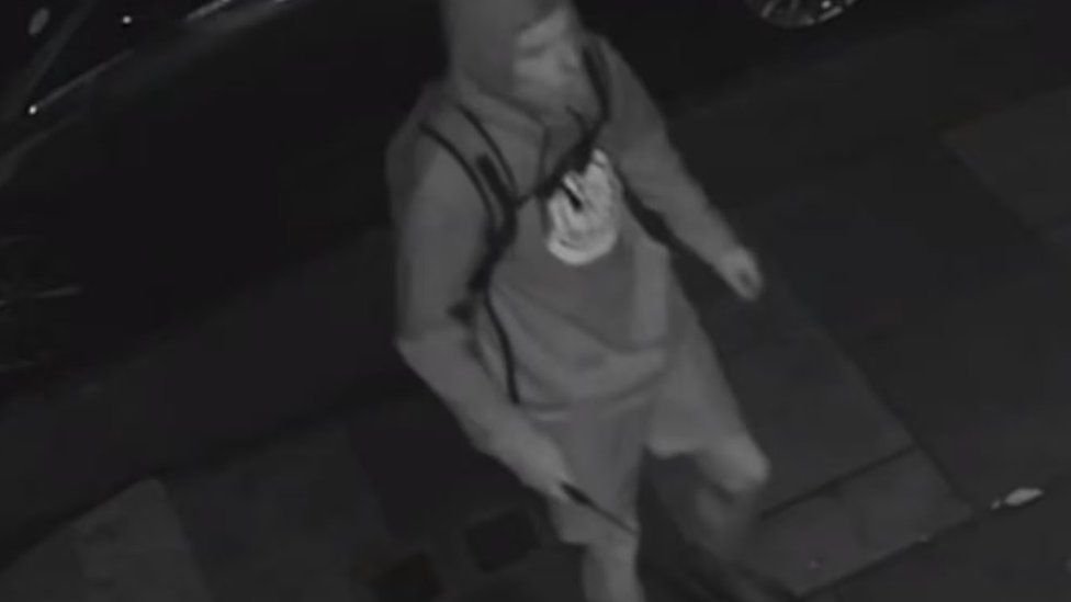 Black and white image of the suspect wearing shorts, a hoodie with a round logo, backpack and trainers