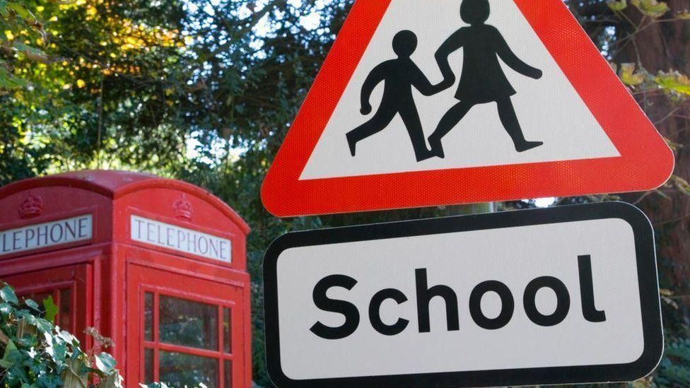 A road sign warning drivers of school children next to a red telephone box