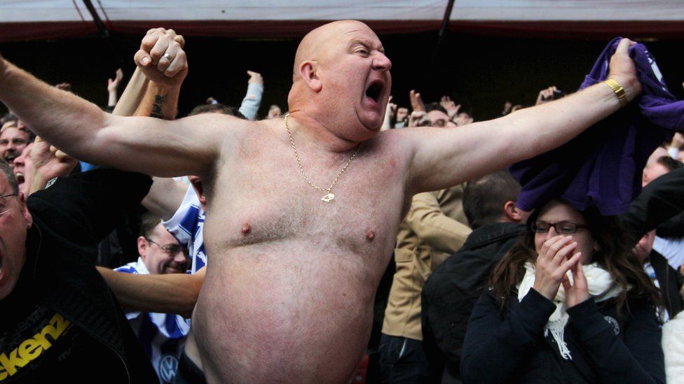 A man shouting, with his shirt off in a crowded football stand