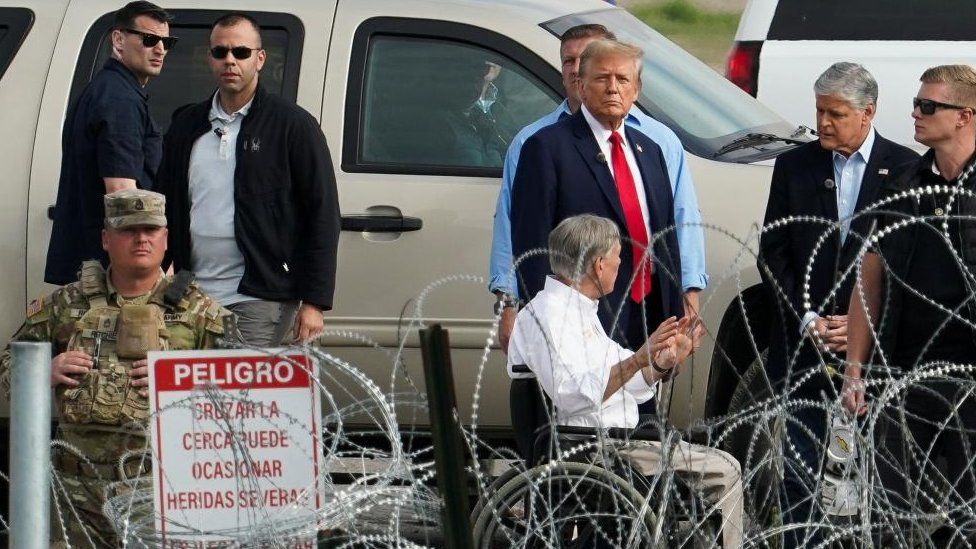 Donald Trump and Greg Abbott pictured alongside border officials, with barbed wire fence in the foreground