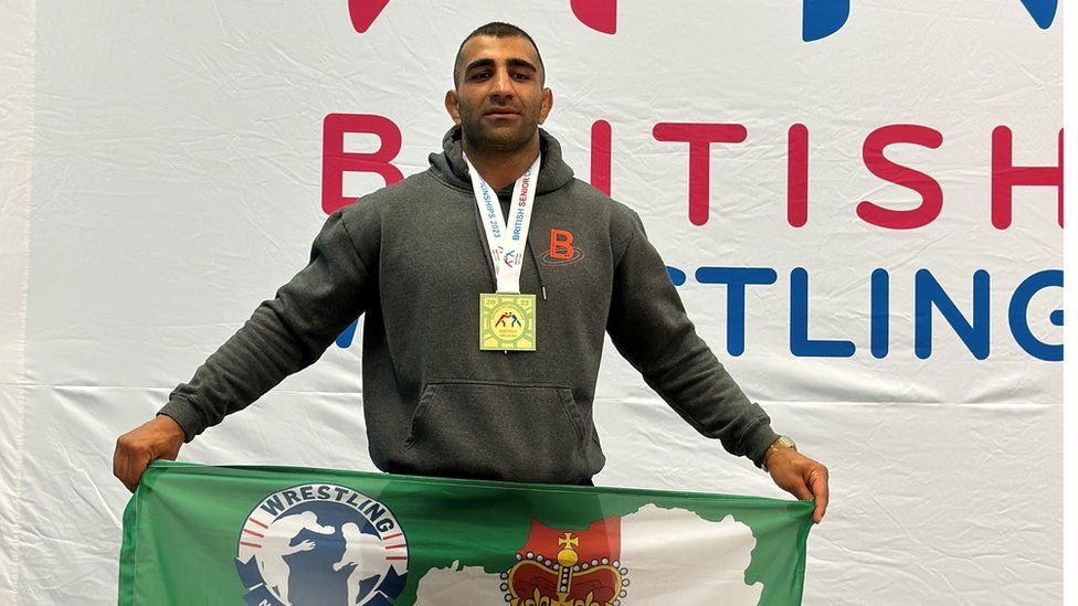 Mehdi Zoodashna poses with a flag after winning a wrestling medal, which is draped around his kneck