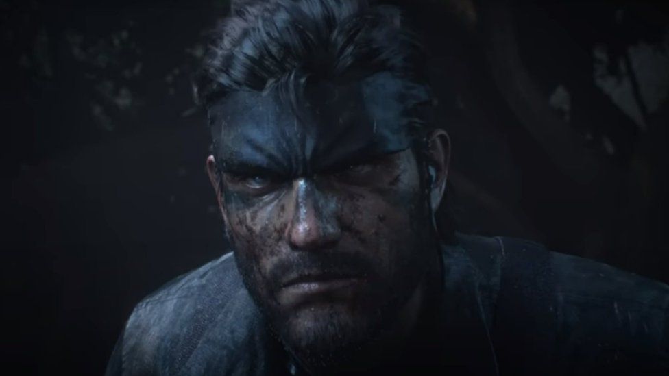 Герой Metal Gear Solid Солид Снейк в джунглях. Он окутан тьмой и с угрожающей хмуростью смотрит вдаль. Его лицо замаскировано грязью, когда он фокусируется на чем-то не в кадре