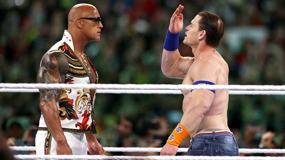 The Rock and John Cena at WrestleMania 40. The Rock is a 51-year-old black man with a muscular physique and tattooed arms and chest. He has a shaved head and wears sunglasses and an embellished white, black, red and gold sleeveless jacket with matching trousers. He stares down John Cena, a 46-year-old white man with brown hair slicked back. John is shirtless, exposing his muscular physique, and wears blue jeans and blue and orange sweat bands on his wrists. He holds his right hand to his head as he faces off with The Rock.