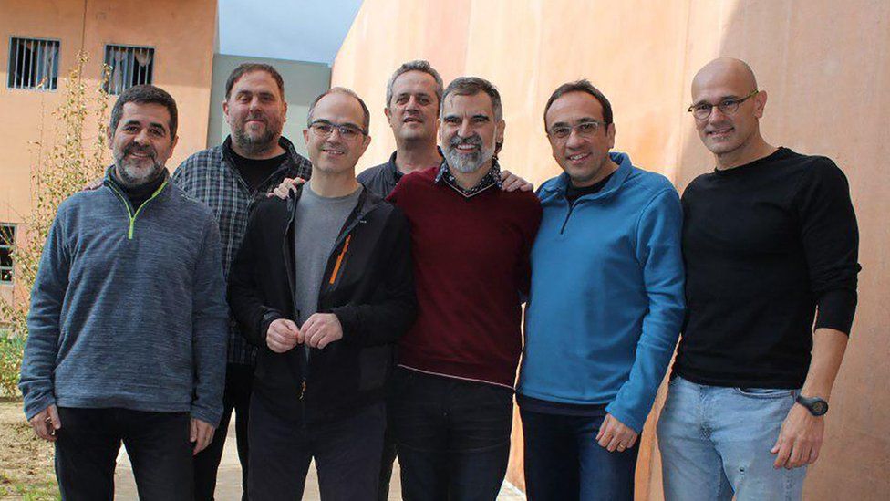 A group photo from a prison courtyard shows seven men, arm in arm, smiling at the camera - from left, they are Jordi Sanchez, Oriol Junqueras, Jordi Turull, Joaquim Forn, Jordi Cuixart, Josep Rull and Raul Romeva