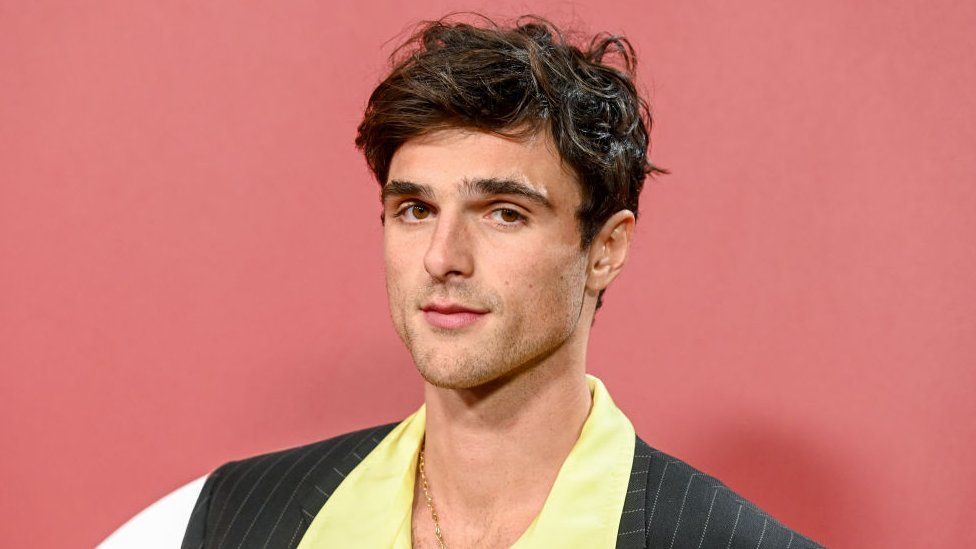 Jacob Elordi pictured at the GQ Men of the Year Party 2023 in Los Angeles. Jacob is a 26-year-old white man with curly brown hair and brown eyes. He is unshaven and wears a grey pinstripe blazer over a lemon yellow shirt with a wide collar. The top buttons are undone revealing a small gold chain. He's pictured in front of a pastel reddish-brown back drop.