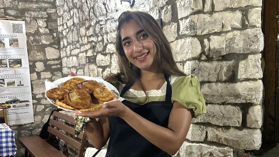 Aliza Ayaz holding a basket of baked pastries. Aliza is a 25-year-old south Asian woman with long brown hair and brown eyes. She wears a black apron over a green short sleeved top and tilts her head towards the basket she's holding at chest height with both hands. She's pictured inside in front of a brick wall with a bench and table in the background