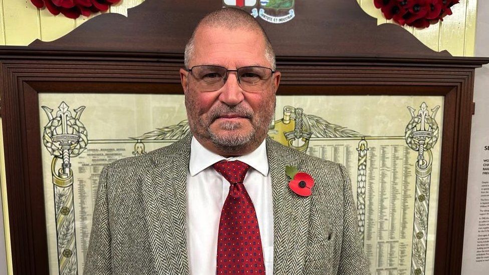 Laurie Hockley. He is wearing glasses and looking directly at the camera. He has very short grey hair and some facial hair. He is wearing a white shirt, red tie and a neutral coloured jacket. There is a poppy pinned on his chest.