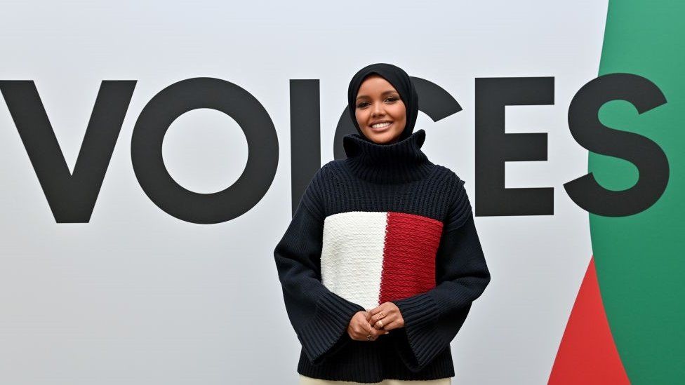 Halima Aden smiling behind a large poster which says: "Voices".