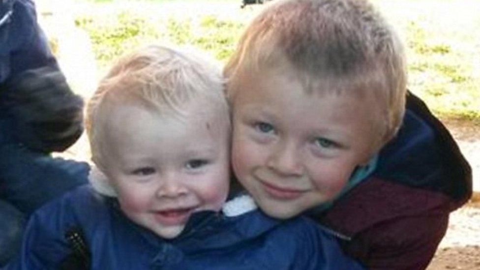 A picture of two young boys, both blonde.
The older boy, wearing a maroon hooded jacket is hugging the younger boy - who is wearing a blue hooded jacket with sheepskin in the hood. 