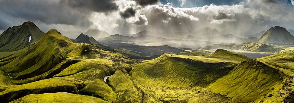 A landscape of green hills with dark clouds and sun breaking through