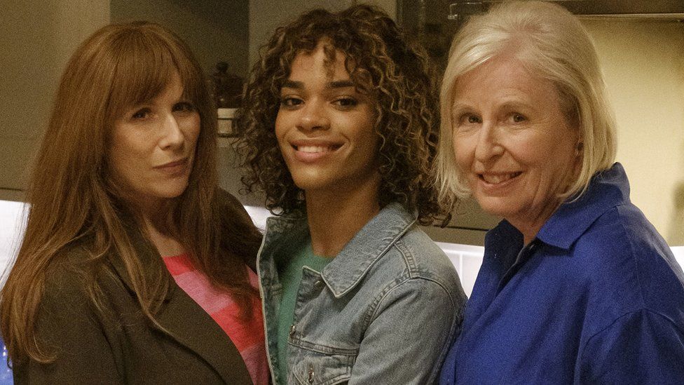 Catherine Tate as Donna, Yasmin Finney as Rose and Jaqueline King as Sylvia in Doctor Who. Donna has long ginger hair with a fringe and wears a dark green blazer over a pink and red striped jumper. Next to her is Rose who has shoulder length curly brown hair and wears a denim jacket over a green jumper. Completing the row is Donna, Rose's grandmother, who has shoulder length blonde hair and wears a blue shirt. The trio are pictured inside, with kitchen cabinets behind them.
