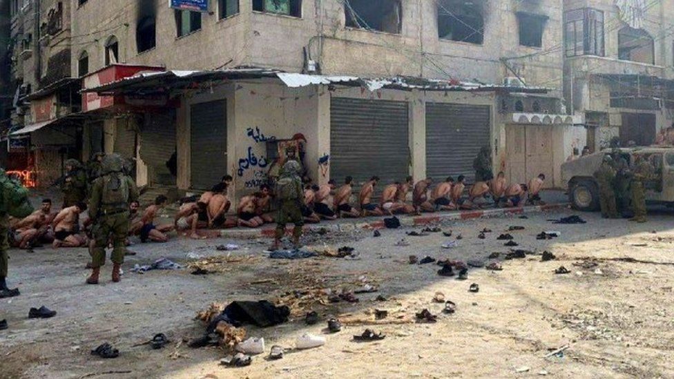 Dozens of men on their knees on a pavement, wearing only underwear, with their hands restrained behind their backs. Israeli soldiers stand guard over them.,