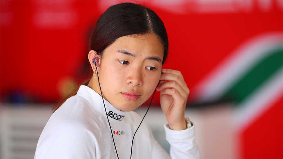 Chloe wearing a white race top, looking down. She is wearing headphones, with her left hand going towards her left ear. The background is blurred but coloured red with a steak of green.