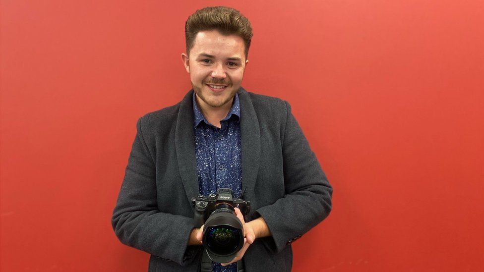 Josh Dury holding a camera and smiling with a red wall behind him