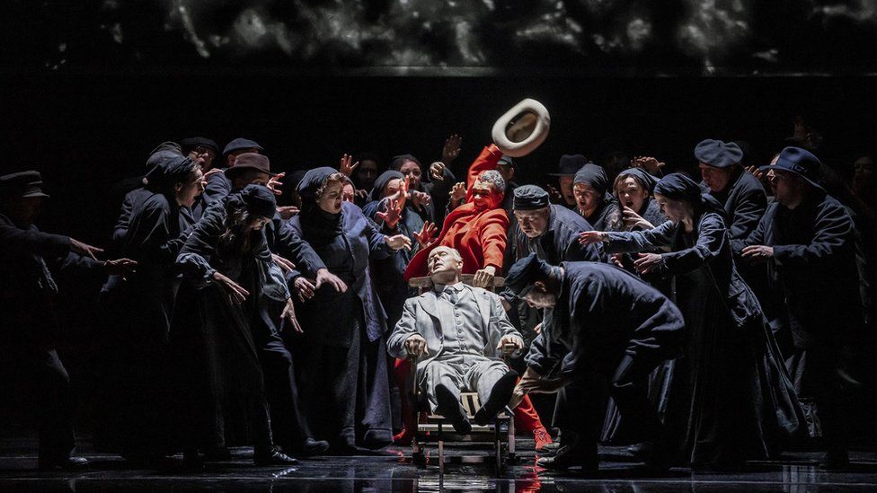 Performers in costume gather round a man lying in a chair on stage at the Bristol Hippodrome