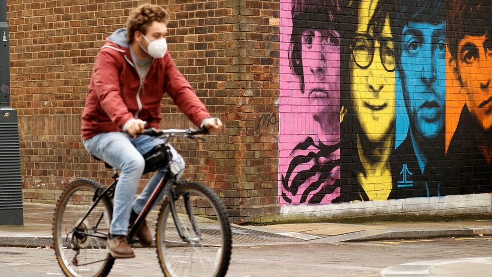 A man wearing a protective mask rides a bike past a mural depicting members of The Beatles in Liverpool