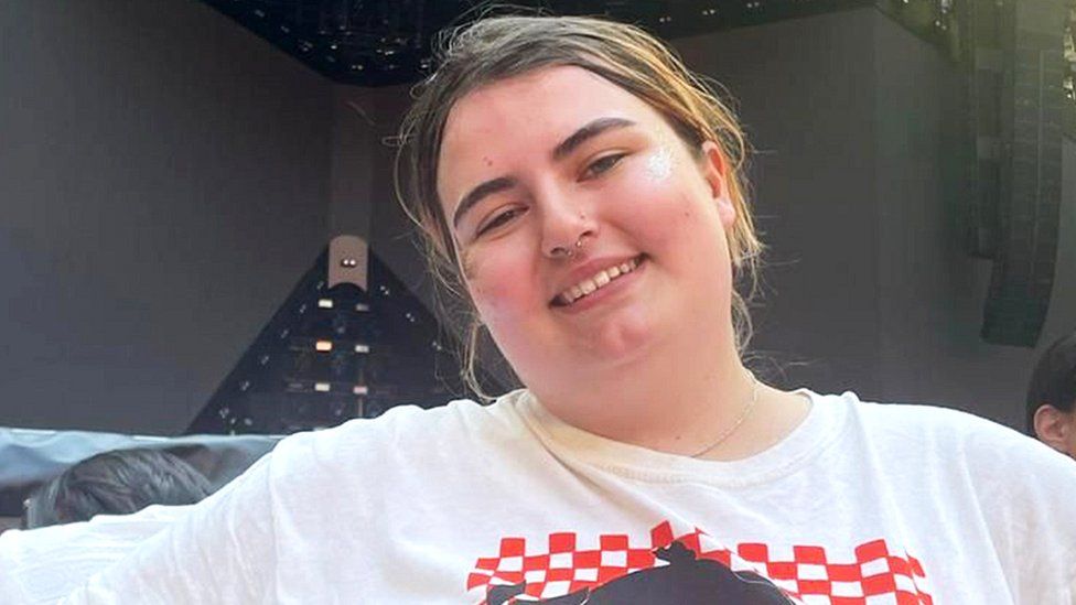 Harriet smiling looking at the camera with her head slightly tilted. She is wearing a white tshirt with red and black squared patterns. The background is of black speaker staging.