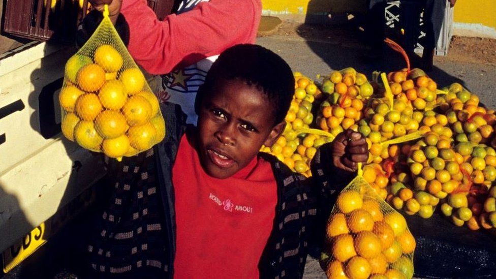 Unidentified children carry oranges from a pickup truck outside Nonqubela train station August 11, 2001 in Site B Khayelitsha, a township located approximately 21 miles outside Cape Town, South Africa.
