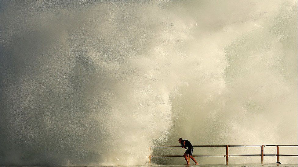 Man grabs onto railing as huge wave splashes against ocean pool