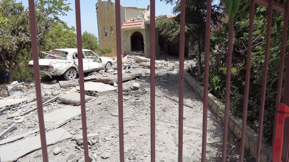 A white car parked outside a destroyed villa locked behind a gate.
