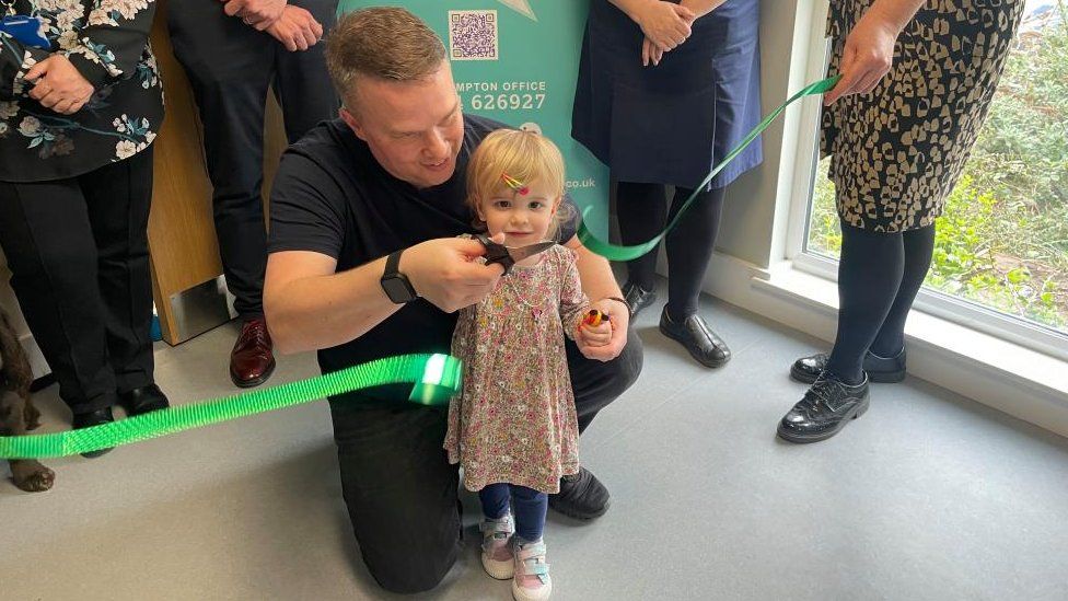 Chloe holds the scissors with her father kneeling beside her, having just cut a green ribbon as part of the hospital's unveiling