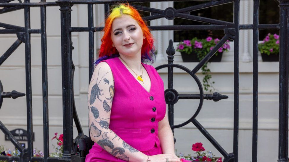 Eliza Rain. Eliza is a 26-year-old white woman with hair dyed yellow, orange, red and purple. She wears a bright pink waistcoat and trousers and a gold necklace. Her right arm has a number of large insect tattoos and she smiles at the camera, looking over her right shoulder. She is pictured outside in front of a white building and iron fence with a number of red and pink flowers around her