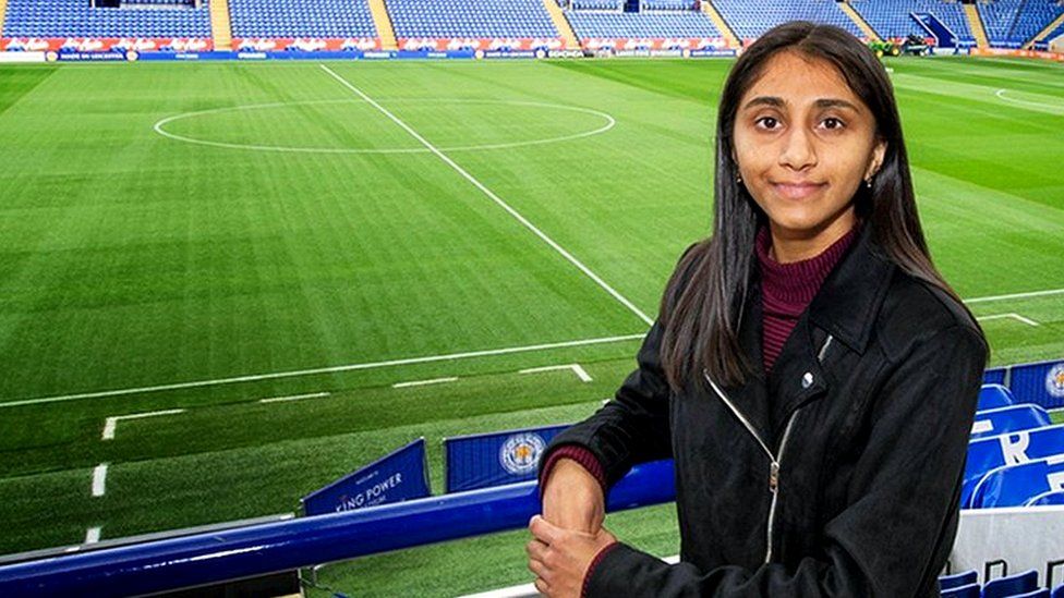 Renuka at Leicester City's stadium. Behind her is the green pitch and empty stands with blue seats, with white writing spelling out Leicester City. Renuka is looking at the camera, leaning on blue railings, with her left hand over her right hand. She is wearing a black jacket with a maroon turtle neck top.