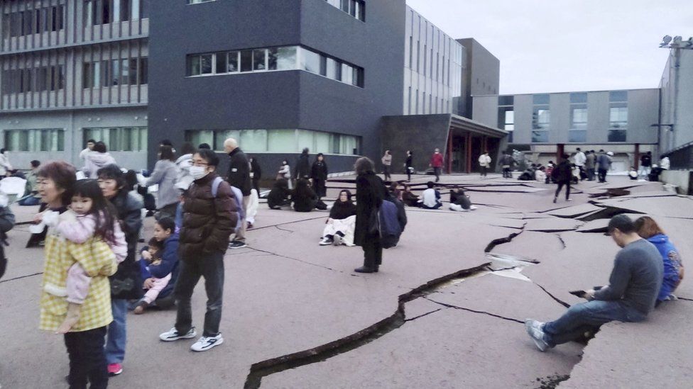 People walk between buildings, on roads and pavements which have split by cracks