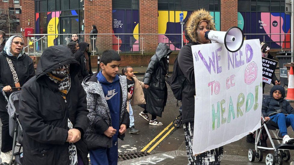 Barton House: Adults and children marching with a woman at the front talking into a megaphone and holding a sign that says 'We need to be heard'