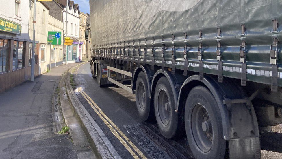A large lorry on a road right next to small shop fronts.