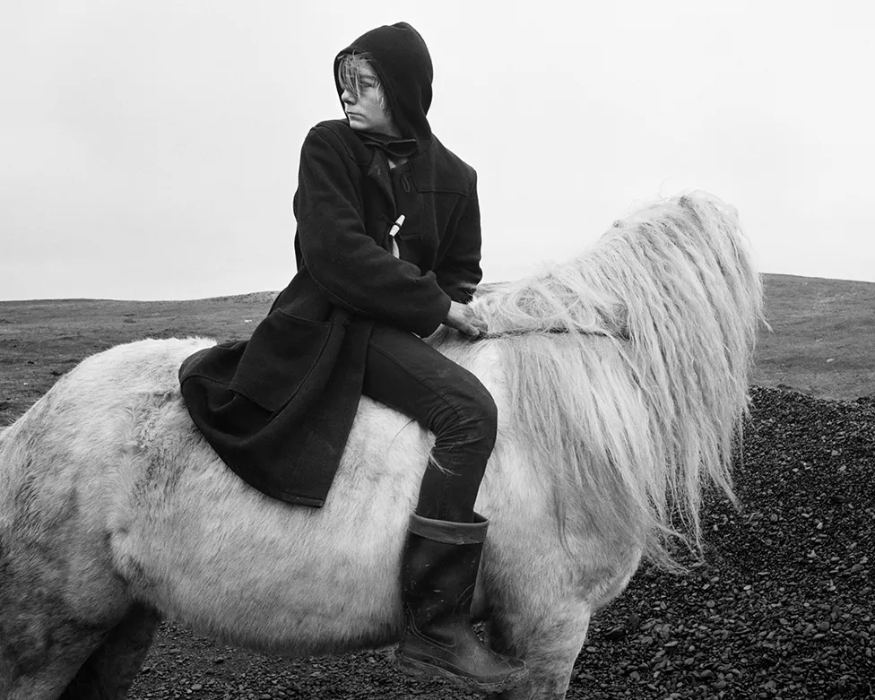 Photo en Noir et Blanc de Chris Killip présentant, sur un cheval blanc de profil, dont la tête est tournée à l'opposée du photographe, monté à cru par une cavalière habillée de noir tournée et regardant vers l'arrière du cheval.