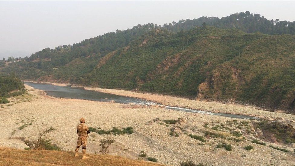 A soldier stands on the rocky shore of a river along the Kashmir Line of Control
